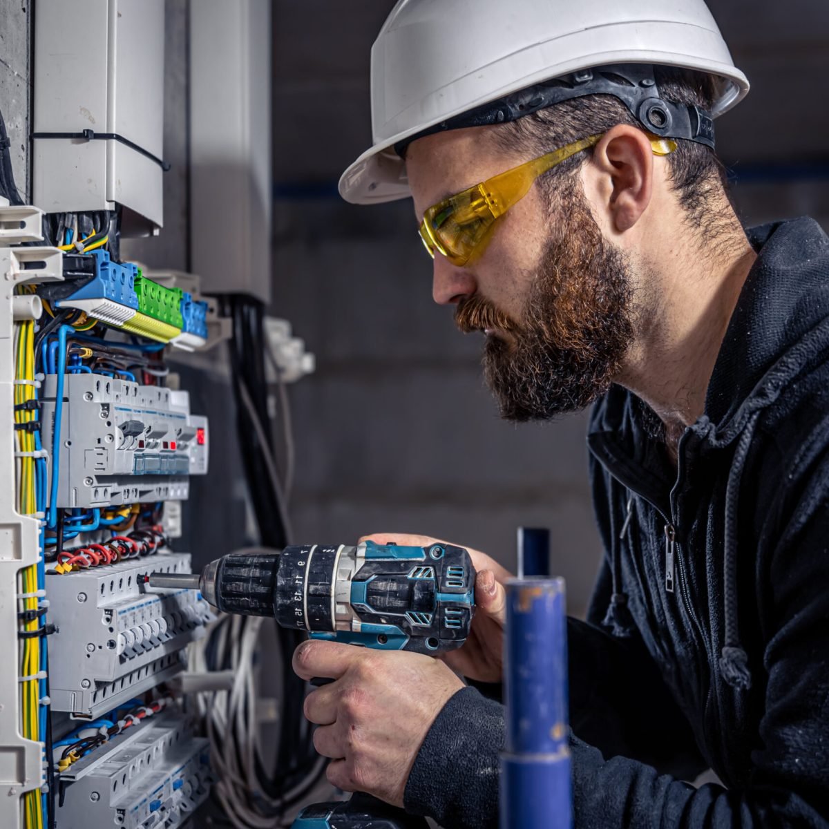 A male electrician works in a switchboard with an electrical connecting cable, connects the equipment with tools.